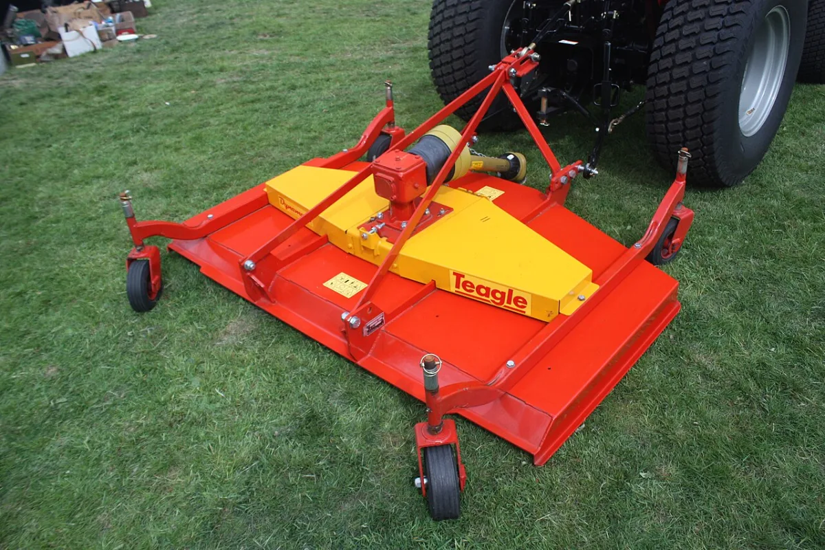 Rood-gele grasmaaier met maaimes gekoppeld aan een tractor, staand op een grasveld